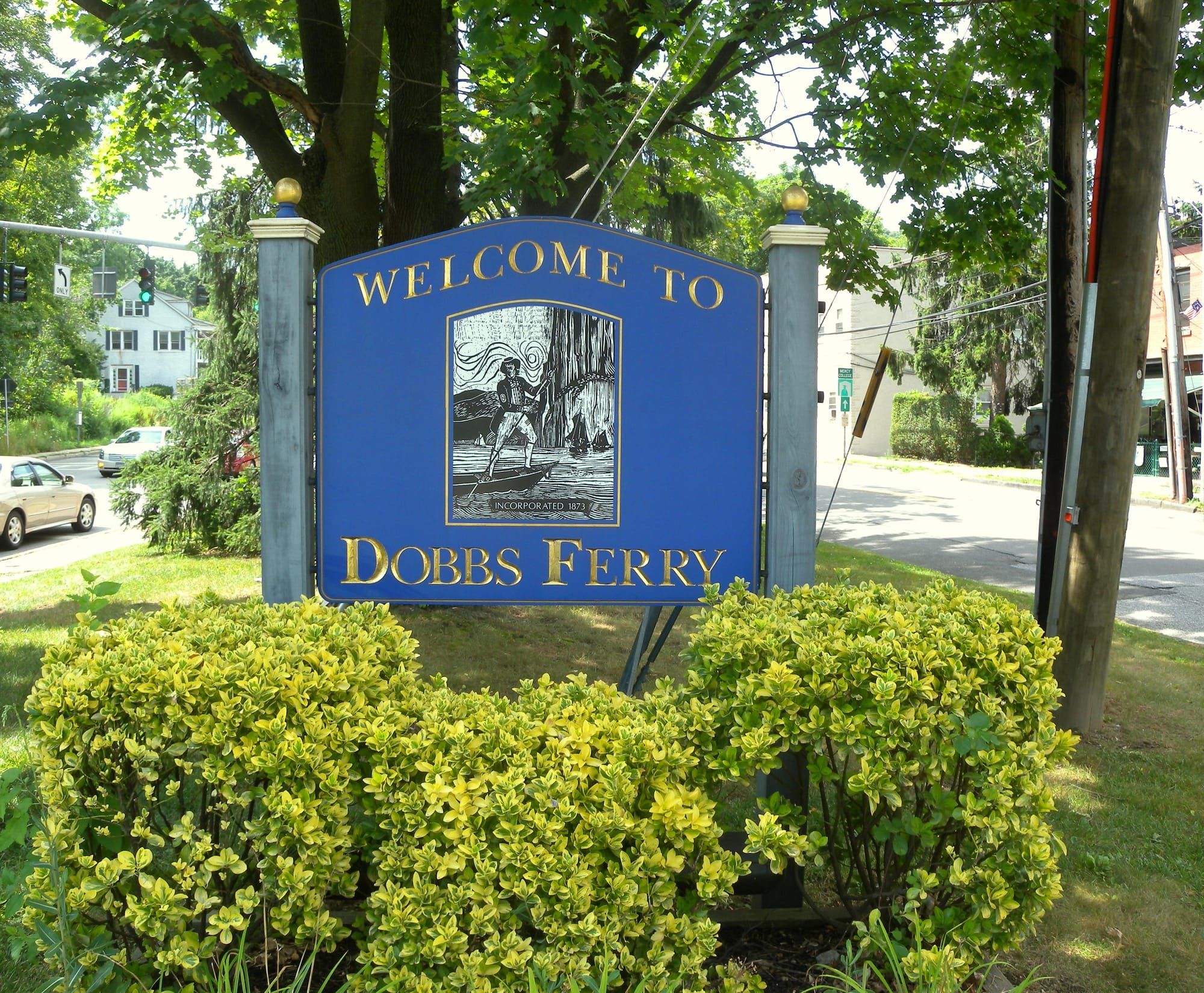 Blue Welcome to Dobbs Ferry sign framed by green shrubs